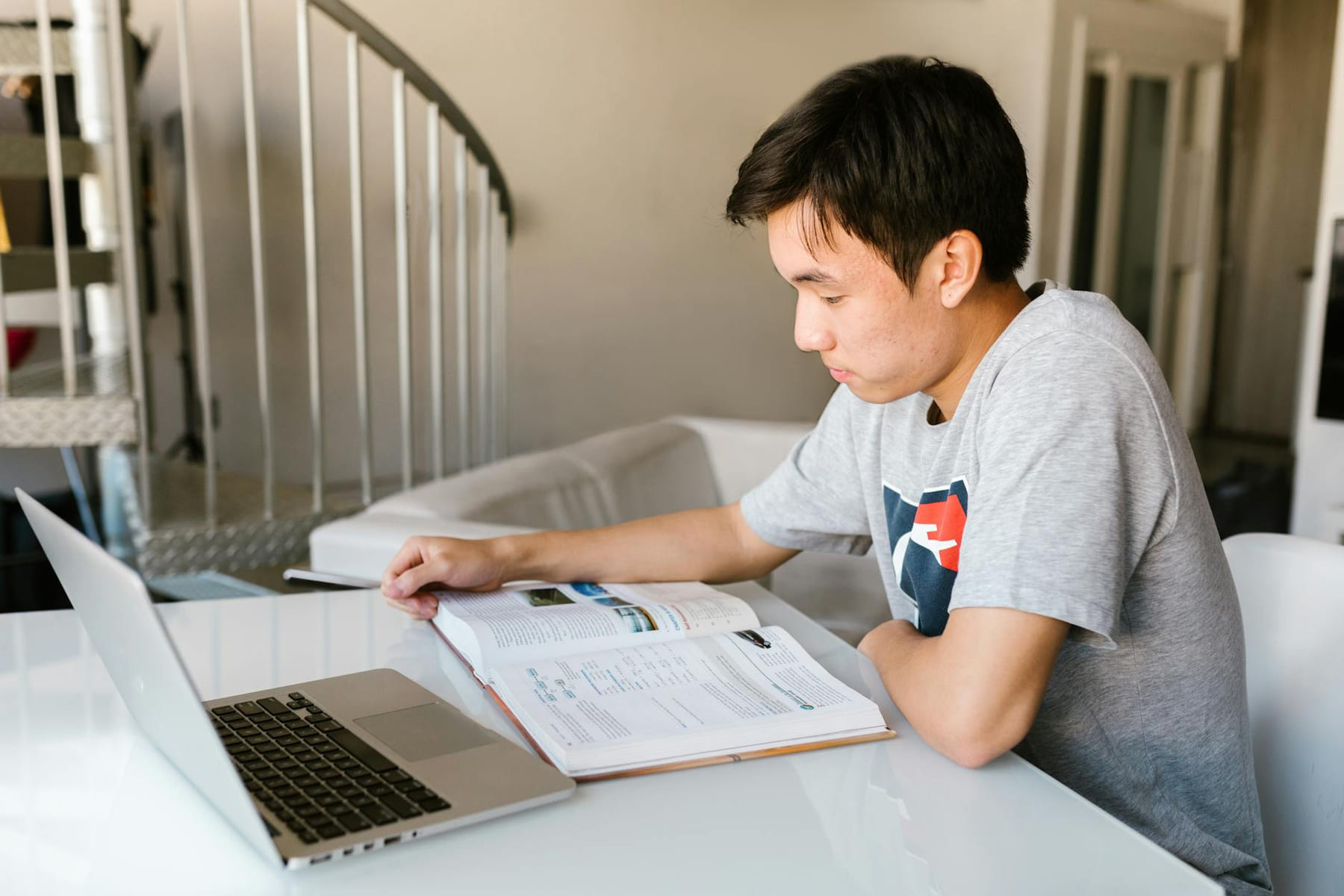 Teenage boy focuses on homework using a laptop and textbook indoors. on Skill Forge