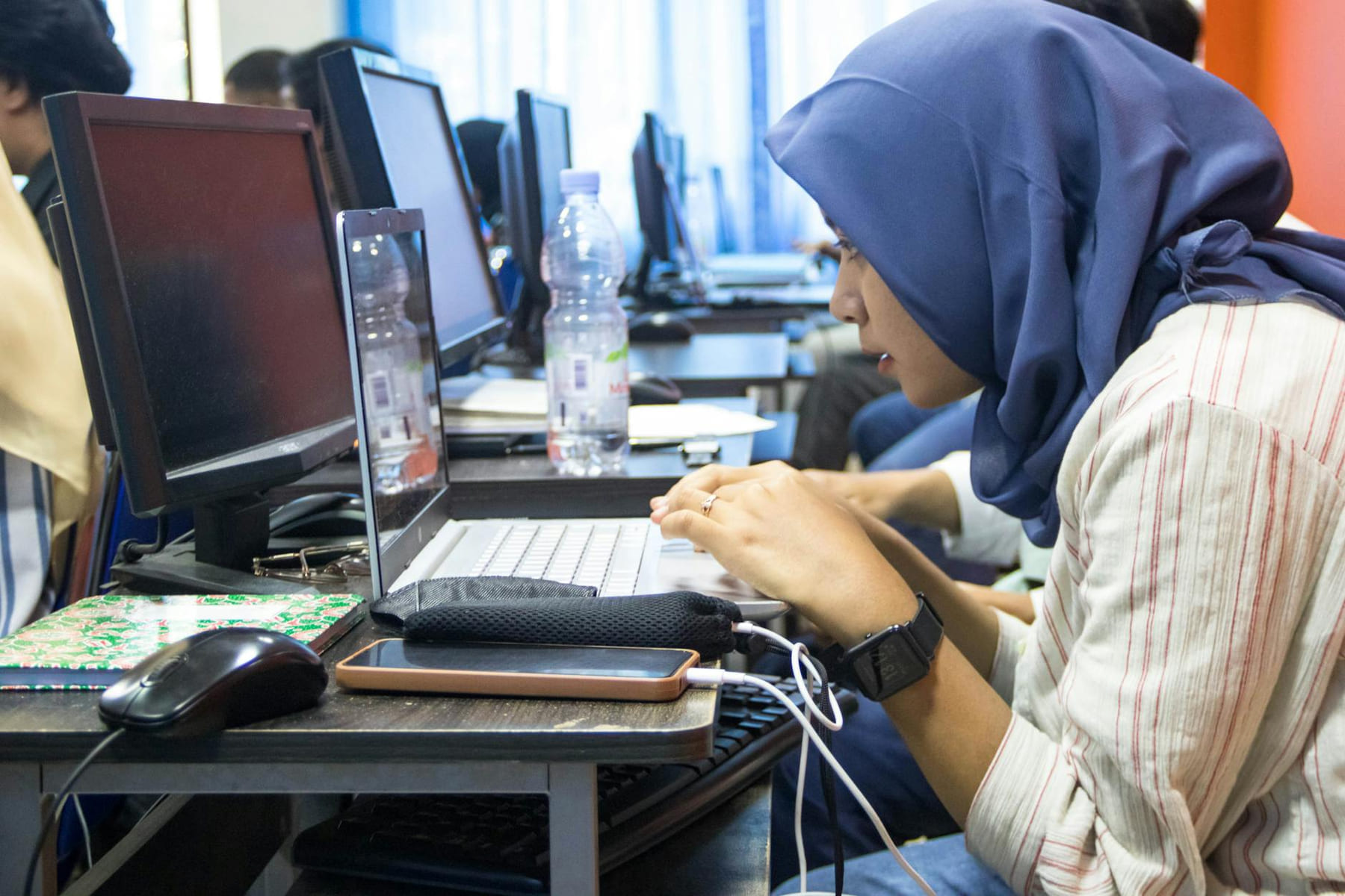 A young woman in hijab using a laptop in a computer classroom setting. on Skill Forge