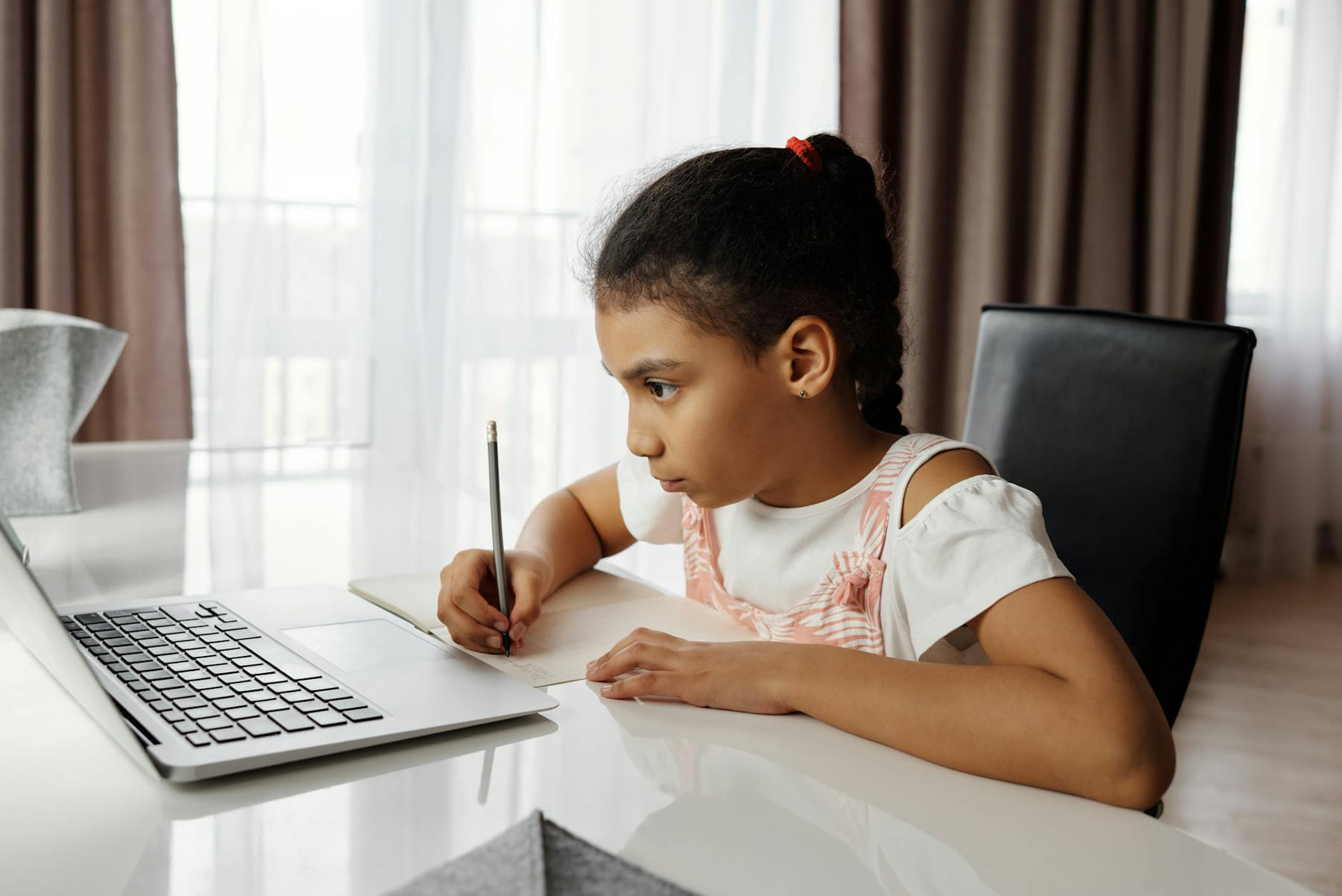 Young girl intensely focused on online learning at home, writing notes by a laptop. on Skill Forge